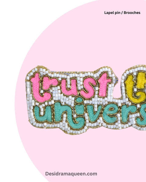 Colorful embroidered lapel pin with the words Trust the Universe, crafted with pink, teal, and yellow beadwork on a white base.