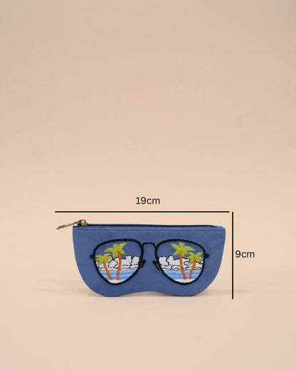 Blue sunglass cover designed in the shape of sunglasses with embroidered tropical palm trees, ocean waves, and cloudy sky, reflecting a serene beach view.