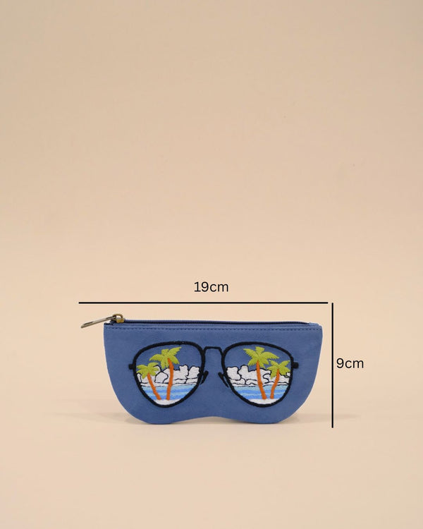 Blue sunglass cover designed in the shape of sunglasses with embroidered tropical palm trees, ocean waves, and cloudy sky, reflecting a serene beach view.