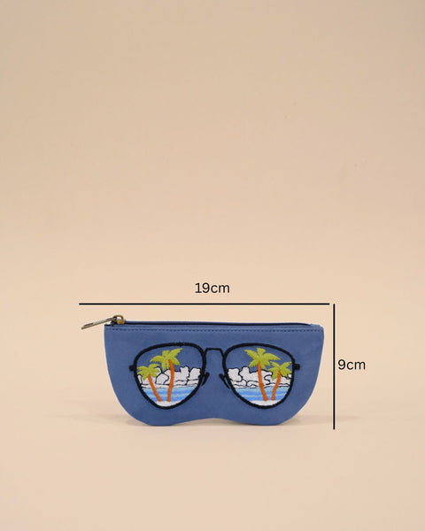 Blue sunglass cover designed in the shape of sunglasses with embroidered tropical palm trees, ocean waves, and cloudy sky, reflecting a serene beach view.