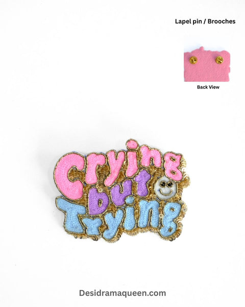 Colorful embroidered brooch with text ‘Crying but Trying’ in pink, purple, and blue with beaded details – handmade quirky statement pin.