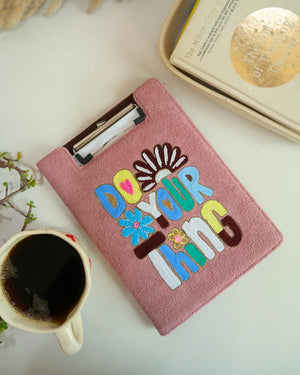 Pink clipboard with colorful text 'Do Your Thing' on a white surface with a coffee cup and books.