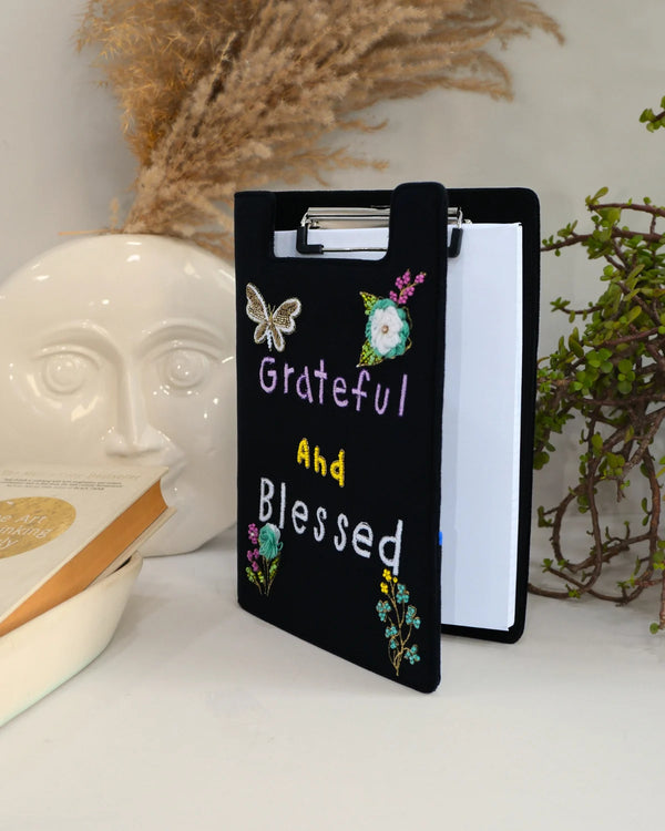 Black clipboard with 'Grateful and Blessed' text and floral designs, placed on a surface with decorative items.