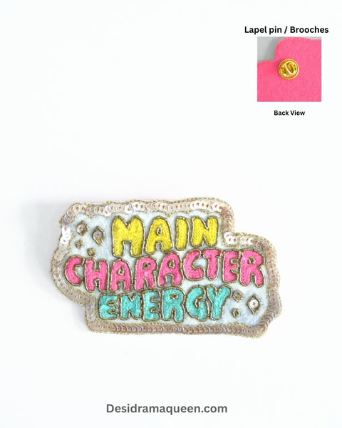 Handmade embroidered brooch with bold text ‘Main Character Energy’ in yellow, pink, and turquoise with shimmering beads.