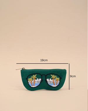 Green fabric sunglass cover designed in the shape of sunglasses with embroidered palm trees, ocean waves, and clouds reflecting a tropical beach view.