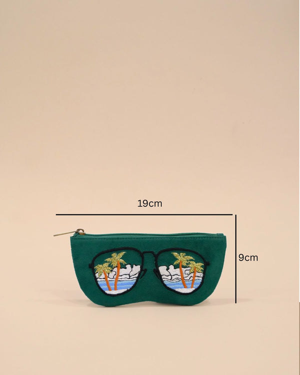 Green fabric sunglass cover designed in the shape of sunglasses with embroidered palm trees, ocean waves, and clouds reflecting a tropical beach view.