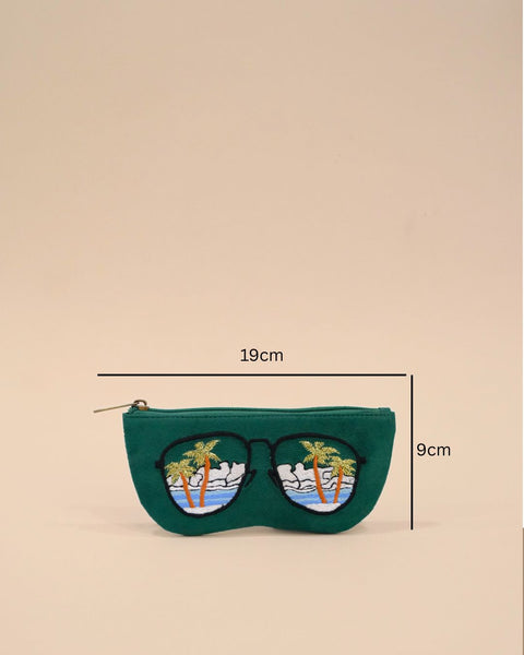 Green fabric sunglass cover designed in the shape of sunglasses with embroidered palm trees, ocean waves, and clouds reflecting a tropical beach view.