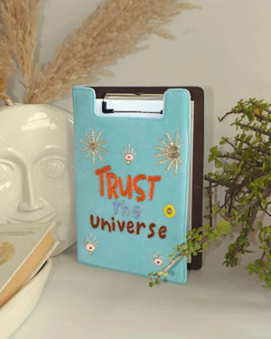 Blue clipboard with 'Trust the Universe' text on a white surface with decorative elements.