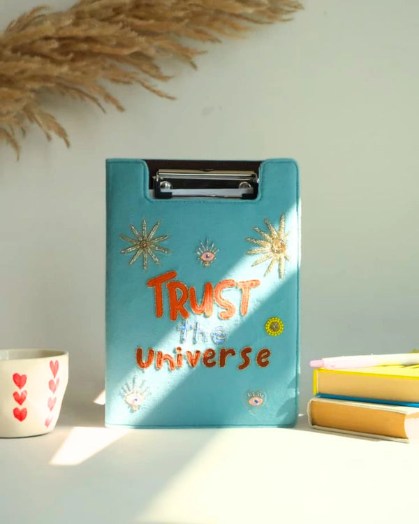 Blue clipboard with 'Trust the Universe' text on a light surface with decorative elements.