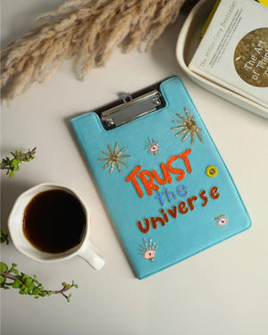 Blue clipboard with 'Trust the Universe' text, coffee cup, and decorative elements on a light surface.