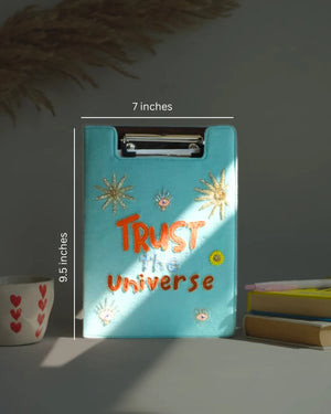Turquoise clipboard with 'Trust the Universe' text, measurements indicated, on a neutral background.