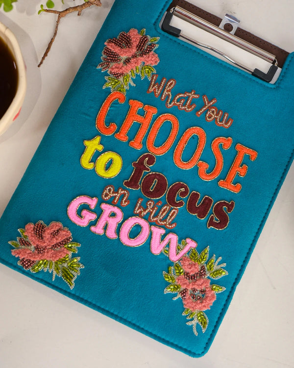 Blue clipboard with motivational quote and floral designs on a white surface.