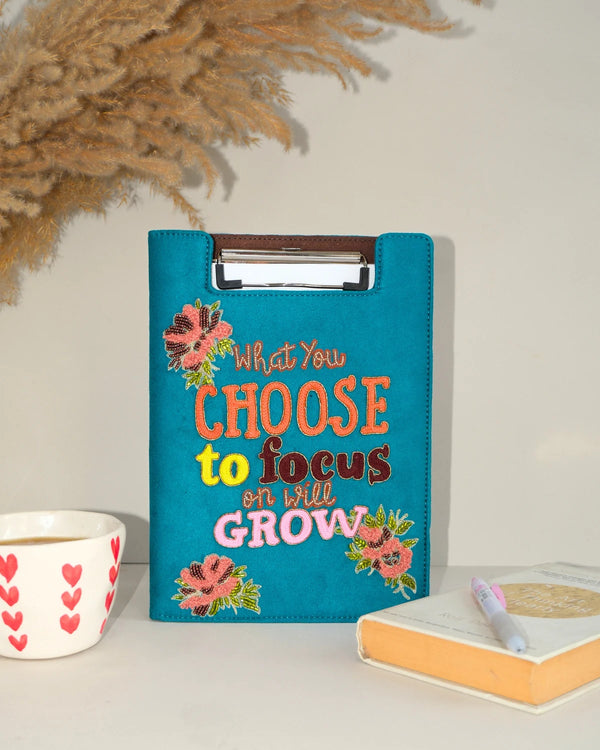 Teal clipboard with motivational quote and floral designs on a surface with decor items.