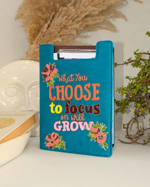 Teal notepad cover with motivational quote and floral designs on a surface.
