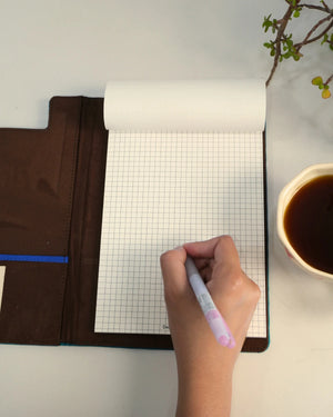 Hand writing on a grid notebook with a cup of coffee and brown bag in the background